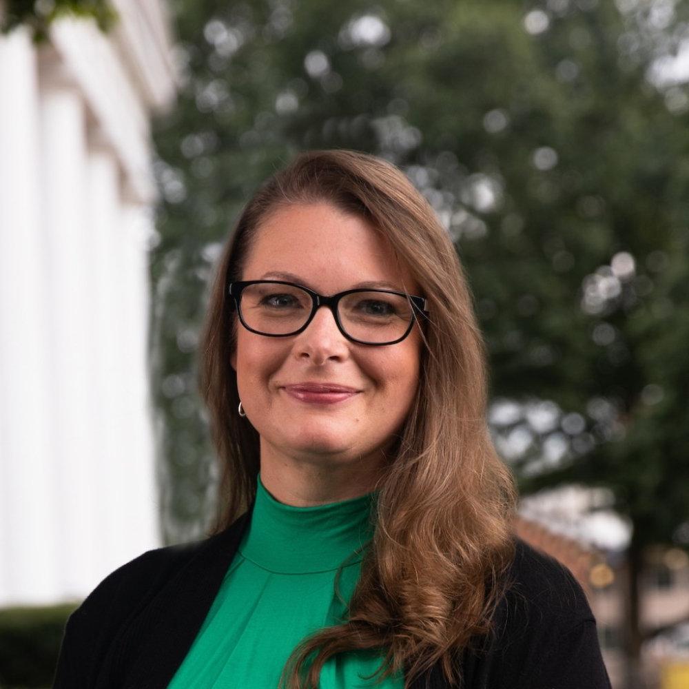 Lady with dark brown hair shoulder length wearing green top and black cardigan with black glasses